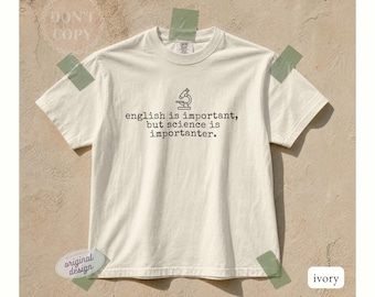 May include: An ivory short-sleeve t-shirt featuring a black line-art illustration of a microscope centered above the text, "english is important, but science is importanter." The shirt has a crew neckline and a standard fit, presented in a neutral tone.