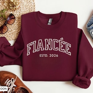 May include: Maroon crewneck sweatshirt with the word "FIANC&Eacute;E" in white block letters, with "ESTD. 2026" underneath. The sweatshirt is displayed with sunglasses, brown shoes, and denim jeans.