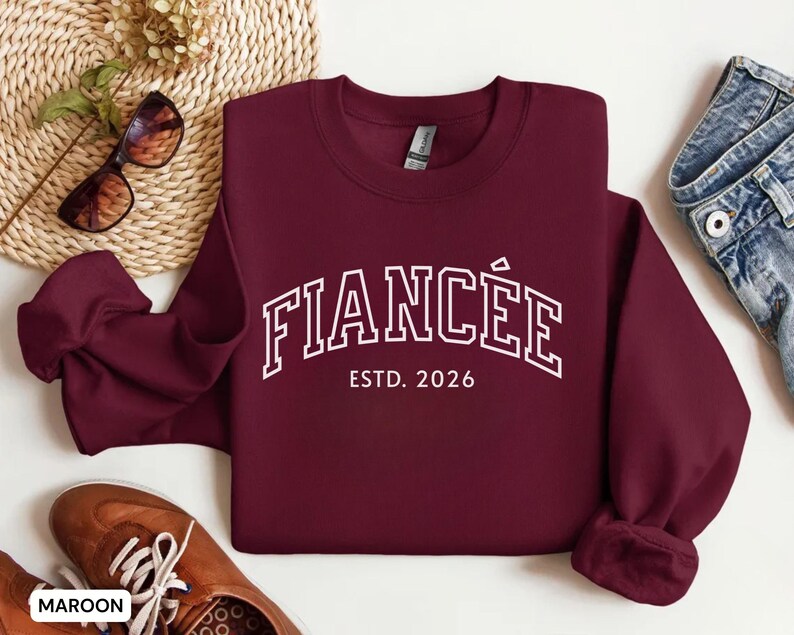May include: Maroon crewneck sweatshirt with the word "FIANC&Eacute;E" in white block letters, with "ESTD. 2026" underneath. The sweatshirt is displayed with sunglasses, brown shoes, and denim jeans.