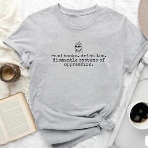 May include: A light gray short-sleeved t-shirt featuring a black line drawing of a cat wearing sunglasses and a party hat, with the text: read books. drink tea. dismantle systems of oppression. The shirt has a crew neckline and a casual fit.