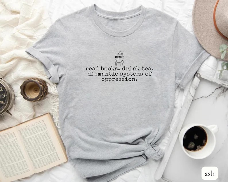 May include: A light gray short-sleeved t-shirt featuring a black line drawing of a cat wearing sunglasses and a party hat, with the text: read books. drink tea. dismantle systems of oppression. The shirt has a crew neckline and a casual fit.