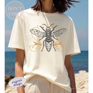 May include: Cream-colored t-shirt featuring a detailed illustration of a bee with outstretched wings. The bee is in black and white, with gold accents and decorative flourishes. The text "Sweet Forever" is below the bee.