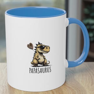 Puede incluir: Taza de cer&aacute;mica blanca con interior y asa azules. La taza presenta un dinosaurio de dibujos animados con gafas de sol y un globo en forma de coraz&oacute;n. La palabra "Papasaurus" est&aacute; impresa debajo.