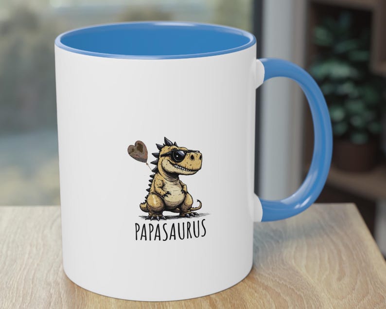 Puede incluir: Taza de cer&aacute;mica blanca con interior y asa azules. La taza presenta un dinosaurio de dibujos animados con gafas de sol y un globo en forma de coraz&oacute;n. La palabra "Papasaurus" est&aacute; impresa debajo.