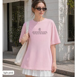 May include: A light pink t-shirt featuring a small graphic of a cat wearing sunglasses and the text: she believed she could, but the cat was asleep on her lap so she didn't. The shirt has a crew neckline and short sleeves, paired with a white skirt.
