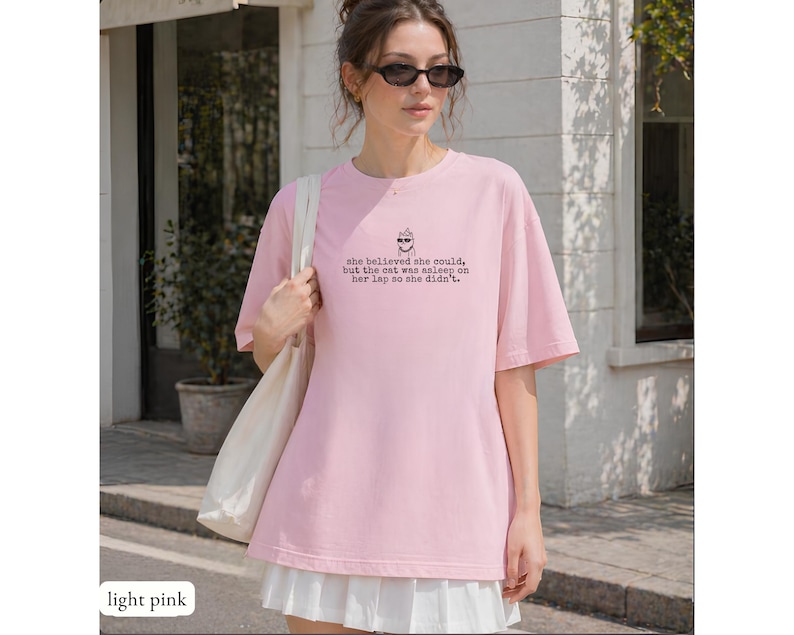 May include: A light pink t-shirt featuring a small graphic of a cat wearing sunglasses and the text: she believed she could, but the cat was asleep on her lap so she didn't. The shirt has a crew neckline and short sleeves, paired with a white skirt.
