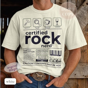 Könnte beinhalten: Ein cremefarbenes Kurzarm-T-Shirt mit dunkelblauem Grafikdruck. Das Design zeigt Symbole für einen Hammer, eine Lupe, einen Stein und ein Glas sowie den Text "certified rock nerd" und "CONTENT DESCRIPTION" neben einem Strichcode.