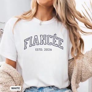 May include: White t-shirt with the word "FIANC&Eacute;E" in navy blue block letters, with "ESTD. 2026" underneath. The shirt is a casual style.