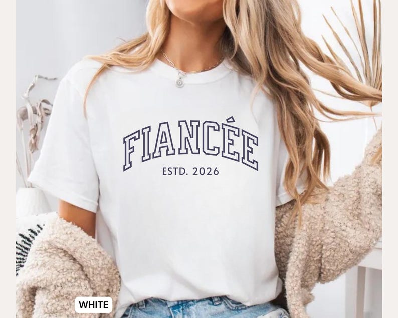 May include: White t-shirt with the word "FIANC&Eacute;E" in navy blue block letters, with "ESTD. 2026" underneath. The shirt is a casual style.