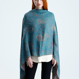Peut inclure: Un châle en paisley turquoise avec un motif détaillé dans des tons orange et marron. Le châle est drapé sur les épaules et a une bordure à franges. Le modèle porte une chemise blanche et un pantalon noir.
