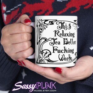 May include: White ceramic mug with black floral art nouveau style design and the text "This Relaxing Tea Better Fucking Work". The mug is held by a person wearing a blue sweater with a red and white pattern.