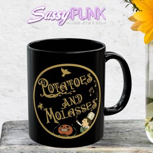 May include: Black ceramic mug featuring a gold-colored design with the words "Potatoes and Molasses" in a circular frame. The design includes illustrations of a bird, musical notes, and a cartoon character. The mug has a curved handle.