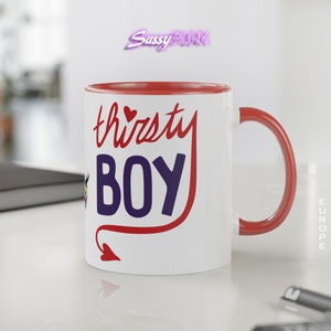 May include: White ceramic mug with a red rim and handle. The mug features a red devil tail with a heart at the end and the text "thirsty BOY" in red and blue lettering.