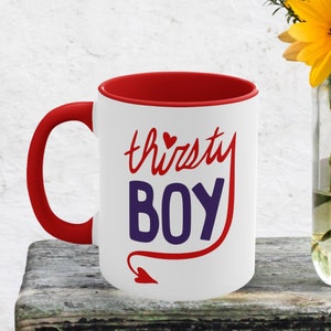 May include: White ceramic mug with a red rim and handle. The mug features a red devil tail with a heart at the end and the text "thirsty BOY" in red and purple.