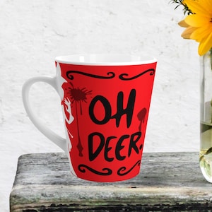 Peut inclure: Une tasse à café en céramique blanche avec un fond rouge et un texte noir qui dit "Oh Deer". La tasse a une poignée blanche et un bord blanc.