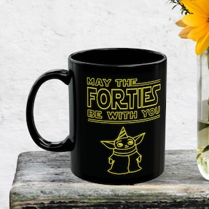 Puede incluir: Taza de café negra con un gráfico amarillo que dice "Que los cuarenta estén contigo" y una imagen de dibujos animados de un personaje de la franquicia Star Wars con un gorro de fiesta.