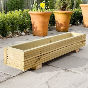 May include: A long, rectangular wooden planter box with a slatted design. The planter is sitting on a patio with stone paving slabs. The planter is empty.