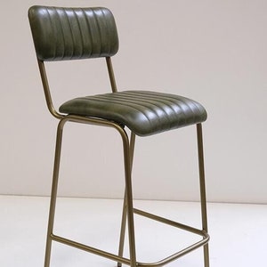 May include: A green leather bar stool with a ribbed seat and backrest. The stool has a gold-colored metal frame and a footrest. The design is a mid-century modern style.