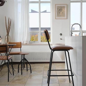 May include: A contemporary kitchen interior with a black metal bar stool. The stool has a brown leather seat and backrest. Other chairs and a table are also visible. The stool is near a white countertop and window.