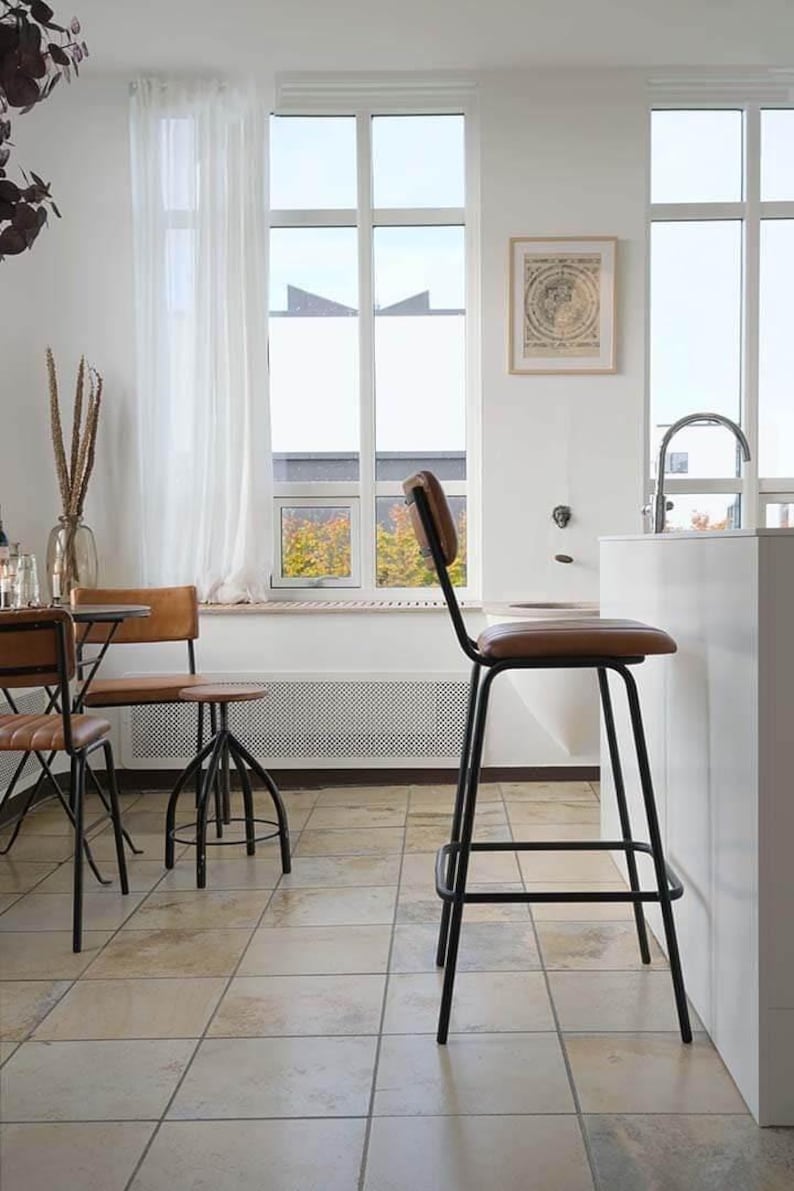May include: A modern kitchen scene featuring a black metal bar stool with a brown leather seat and backrest. The stool is positioned near a white countertop and window. Other chairs and a table are also visible.