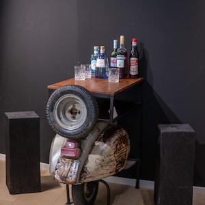 Könnte beinhalten: Ein origineller Bartisch aus einem Vintage-Roller, mit einer Holzplatte und einem Ersatzreifen. Der Wagen enthält mehrere Flaschen Spirituosen und Gläser, flankiert von zwei dunklen Holzblockhockern. Der Roller ist in einem verwitterten Off-White lackiert.