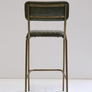 May include: A tall bar stool with a green, padded seat and backrest. The frame is made of gold-colored metal. The backrest has horizontal metal accents. The stool's height is suitable for a bar or counter.