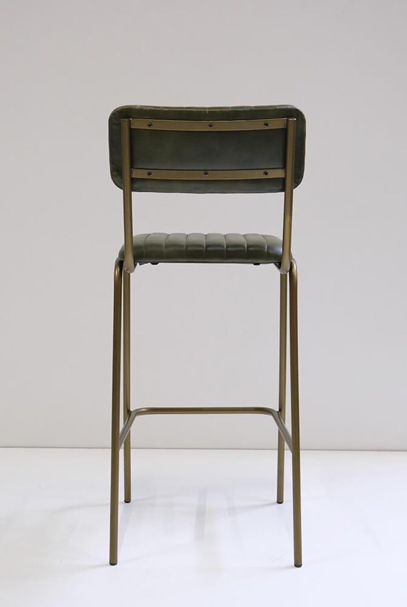 May include: A tall bar stool with a green, padded seat and backrest. The frame is made of gold-colored metal. The backrest has horizontal metal accents. The stool's height is suitable for a bar or counter.