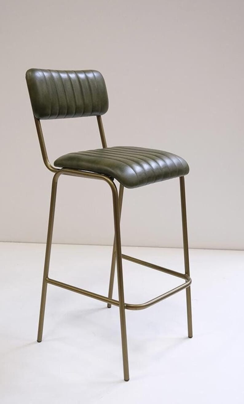 May include: A green leather bar stool with a ribbed seat and backrest. The stool has a gold-colored metal frame and a footrest. The design is a mid-century modern style.