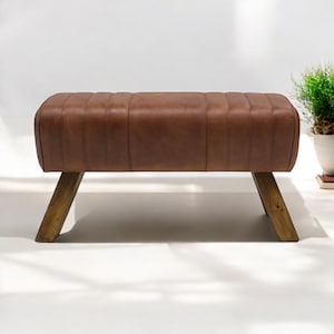 Op de afbeelding: Een bruine leren bank met houten poten. De bank heeft een licht afgeronde bovenkant en een gestoffeerd ontwerp.