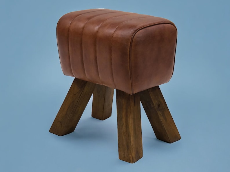 Puede incluir: Un taburete de cuero marr&oacute;n y madera con la parte superior redondeada y cuatro patas. El taburete est&aacute; hecho de madera marr&oacute;n oscura y tiene un asiento de cuero marr&oacute;n.