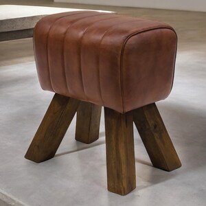 Puede incluir: Un taburete tapizado en cuero marr&oacute;n con cuatro patas de madera. El taburete tiene una forma rectangular y una parte superior ligeramente redondeada.