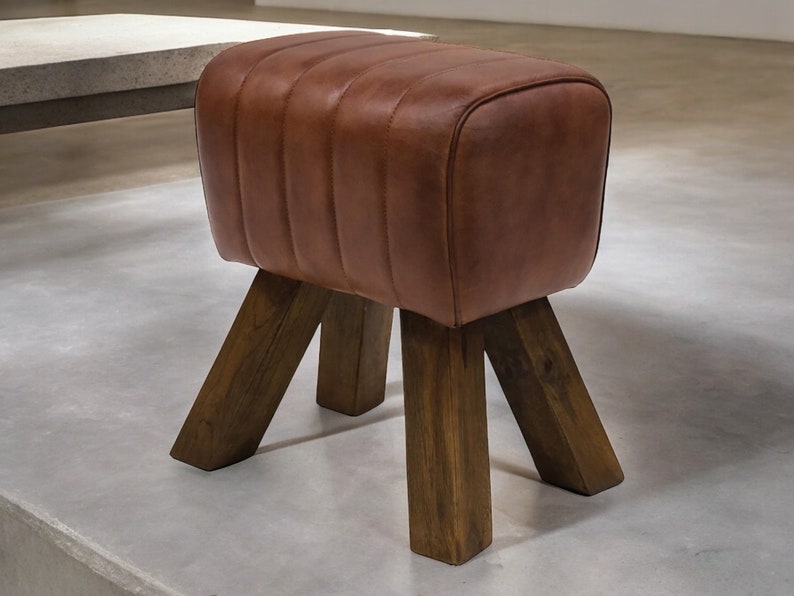 Puede incluir: Un taburete tapizado en cuero marr&oacute;n con cuatro patas de madera. El taburete tiene una forma rectangular y una parte superior ligeramente redondeada.