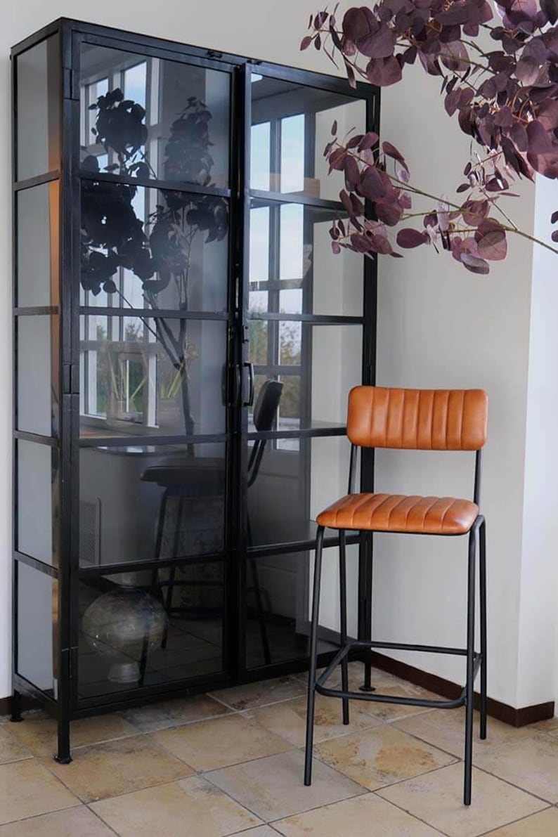 May include: A black metal framed display cabinet with glass panels, and a brown leather and black metal bar stool. The cabinet has a dark interior with a vase and a stool visible inside. Purple foliage is in the background.