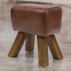 Puede incluir: Un taburete de caballo de gimnasia de cuero marr&oacute;n con cuatro patas de madera. El taburete est&aacute; hecho de madera maciza y tiene una construcci&oacute;n robusta.