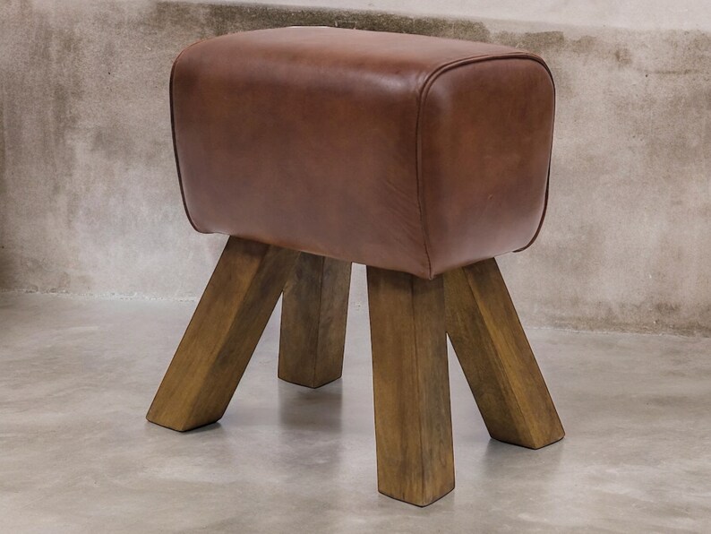 Puede incluir: Un taburete de caballo de gimnasia de cuero marr&oacute;n con cuatro patas de madera. El taburete est&aacute; hecho de madera maciza y tiene una construcci&oacute;n robusta.