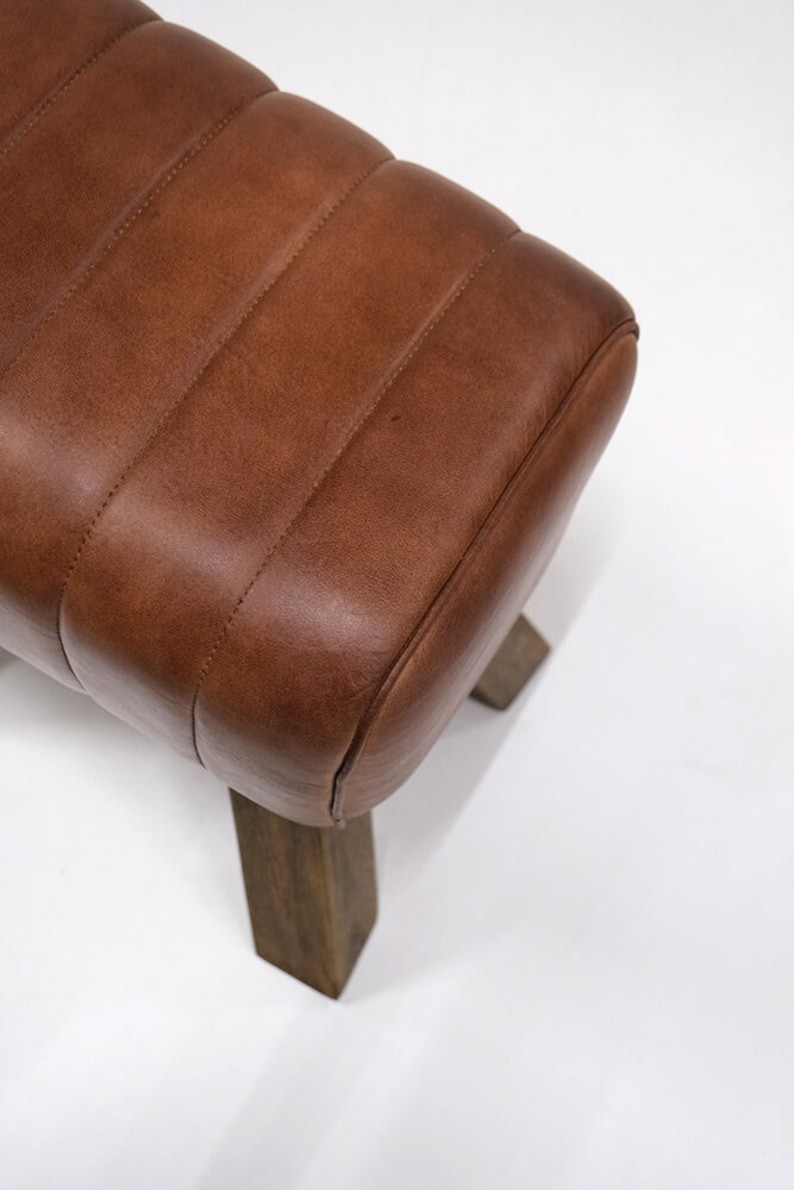 Puede incluir: Un banco de cuero marr&oacute;n con la parte superior redondeada y cuatro patas de madera. El banco tiene un dise&ntilde;o capiton&eacute; y est&aacute; hecho de materiales de alta calidad.