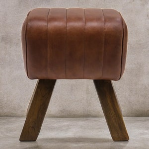 Puede incluir: Un otomano de cuero marrón con una parte superior capitoné y cuatro patas de madera. El otomano está sobre un suelo de hormigón gris.