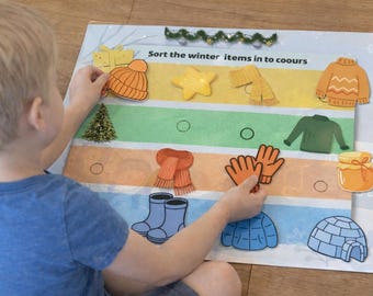 May include: A child sorts winter-themed cutouts on a colorful board. The board features items like a hat, scarf, gloves, and boots. The text "Sort the winter items in to colours" is visible. The child is wearing a blue shirt.
