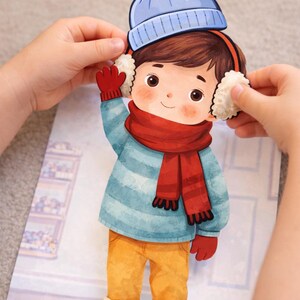 May include: A paper cutout of a child dressed for winter. The child wears a blue hat, earmuffs, a red scarf and gloves, a blue and white striped sweater, yellow trousers, and blue boots. The cutout is held by two hands.
