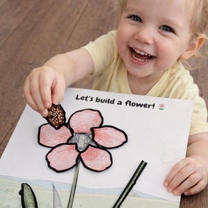 May include: A child is smiling while holding a piece of a flower puzzle. The puzzle is on a white paper with the text "Let's build a flower!". The flower has pink petals and a gray center. The child is wearing a yellow shirt.