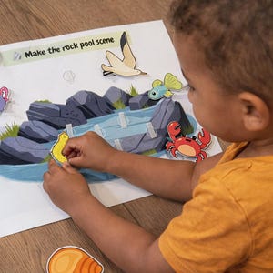 May include: A child is shown interacting with a colorful educational activity. The activity features a rock pool scene with various sea creatures and the text "Make the rock pool scene". The child is placing stickers of sea creatures on the scene.