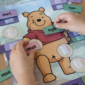Puede incluir: Un juego educativo con Winnie the Pooh. La imagen muestra a un niño emparejando palabras como "ojos" y "pequeño" con las partes correspondientes de una ilustración de Winnie the Pooh. El juego está sobre un fondo azul claro.