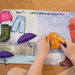 Peut inclure: Un livre éducatif pour enfants ouvert sur deux pages. L'une montre un pull rose, un jean bleu, des bottes vertes et un parapluie violet. L'autre montre une veste jaune, une écharpe rouge et un bonnet rouge. Le texte dit : "Rain - which clothes go where?"