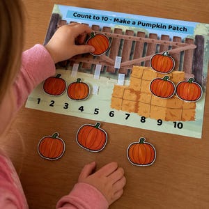 May include: A child's educational game titled "Count to 10 - Make a Pumpkin Patch." The game features a colorful illustration of a pumpkin patch with hay bales and a fence. The child is placing orange pumpkin cutouts onto the game board, learning to count.