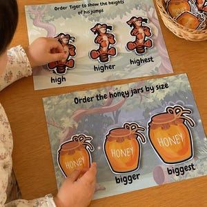 Puede incluir: Juego educativo para niños con Tigger y tarros de miel. El juego incluye tarjetas con Tigger en diferentes alturas de salto y tarros de miel de varios tamaños. El texto en las tarjetas dice "high", "higher", "highest", "bigger" y "biggest".