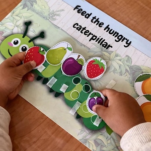 Puede incluir: Juego educativo infantil con una oruga verde y recortes de frutas, como fresas, peras y ciruelas. El texto "Feed the hungry caterpillar" se muestra en el tablero. El juego está diseñado para niños pequeños.