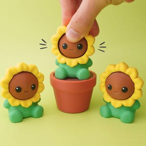 May include: A hand places a yellow sunflower figure into a terracotta pot, with two other similar figures nearby. The figures have brown faces, yellow petal-like heads, and green bodies. The background is a solid light green.
