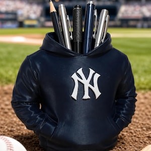 May include: A navy blue pencil holder shaped like a hooded sweatshirt featuring the white New York Yankees logo on the front. The holder contains several pens and a pencil, with a baseball and two wooden bats visible in the foreground.