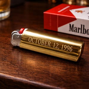 K&ouml;nnte beinhalten: Ein goldfarbenes Feuerzeug mit der Gravur "OCTOBER 12, 1996" liegt auf einer Holzoberfl&auml;che. Eine Packung Marlboro-Zigaretten befindet sich im Hintergrund. Das Feuerzeug hat einen roten Akzent in der N&auml;he der Z&uuml;ndung.