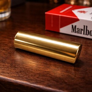 K&ouml;nnte beinhalten: Ein zylindrisches, goldfarbenes Metallobjekt liegt auf einer dunklen Holzoberfl&auml;che. Eine Packung Marlboro-Zigaretten mit rotem und wei&szlig;em Design befindet sich im Hintergrund. Die Szene ist mit warmem Licht beleuchtet.